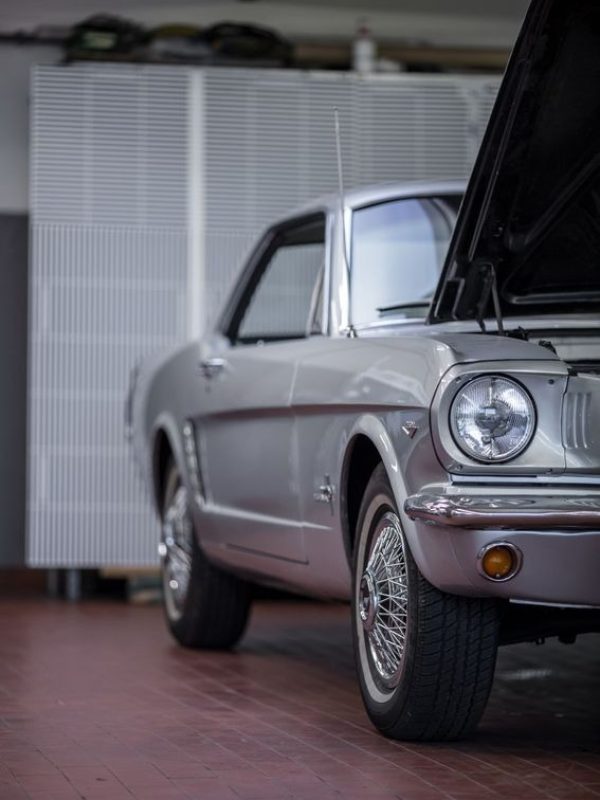 Ein silberner Oldtimer mit offener Motorhaube, geparkt in einem Innenraum auf rotem Fliesenboden.
