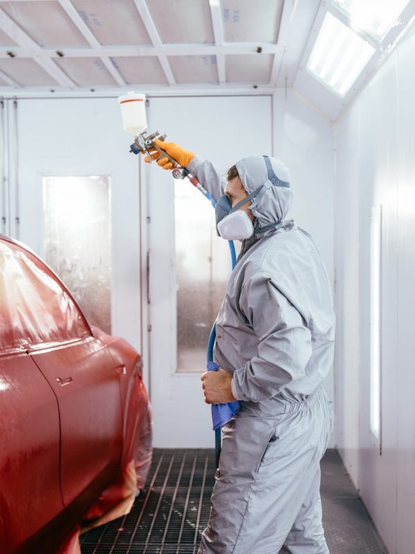 Person in Schutzkleidung bei der Fahrzeuglackierung an einem roten Auto in einer Lackierkabine.