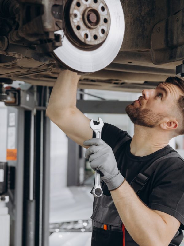 Mechaniker bei der Kfz-Reparatur, der in einer Werkstatt den Unterboden eines Fahrzeugs inspiziert.