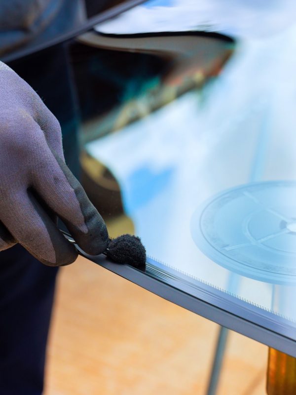 Eine behandschuhte Hand trägt Dichtungsmasse auf den Rand einer großen Glasscheibe auf.